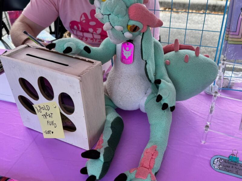A plush kobold wearing a Service Kobold dog tag, and next to a wooden box with a paw print on it that reads Kobold Treat Fund